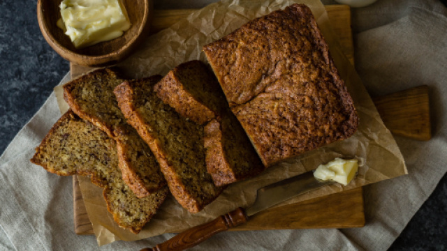 Sliced loaf of banana bread sitting on a cutting board with butter, milk, and a knife di W. Wright