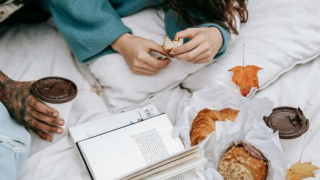 Ragazza che mangia un croissant sul letto con libro- &copy; Pexels